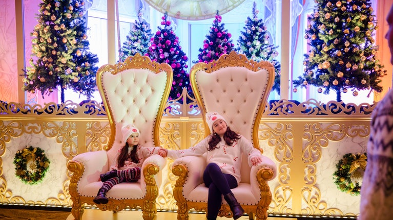Two children in cream and red Christmas jumpers sitting on huge cream and gold thrones with Christmas trees behind, in Mottisfont's Whistler room for A Cinderella Christmas